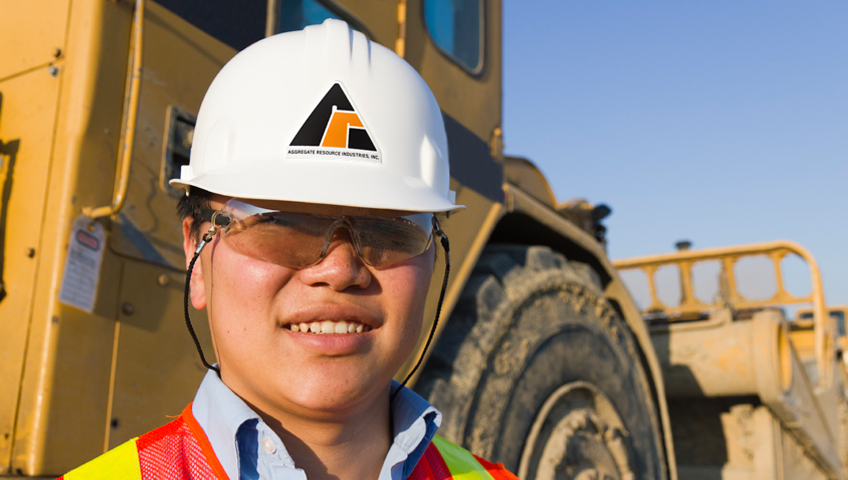 Person wearing construction helmet with ARI logo standing in front of heavy equipment