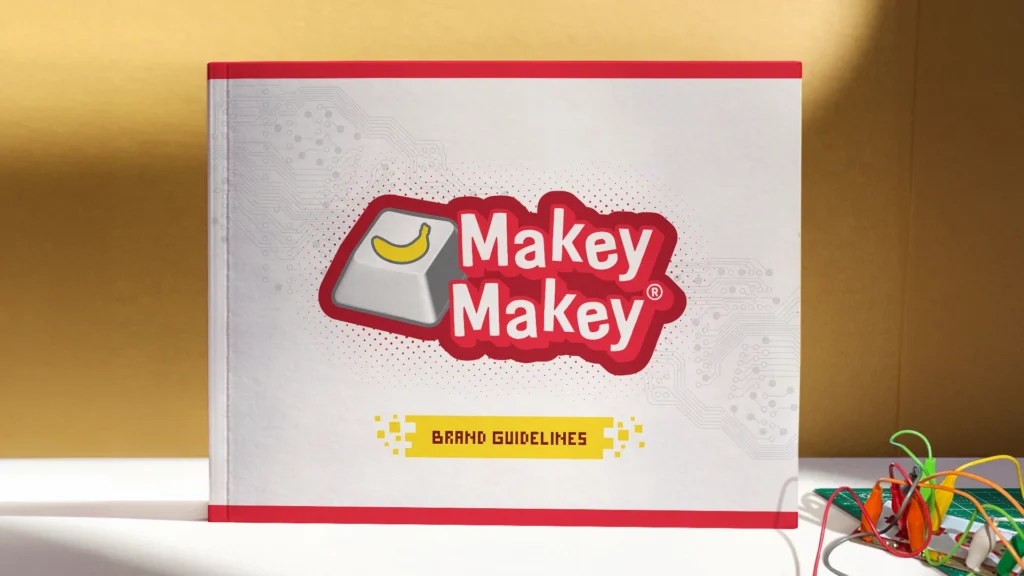 Cover of Makey Makey's Brand Guidelines book with Makey Makey and wires sitting on table